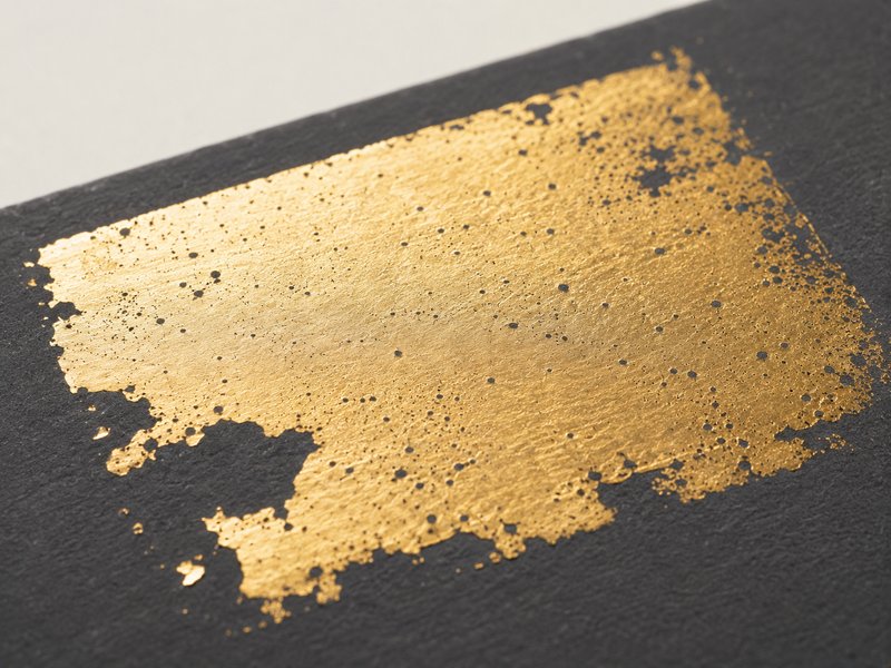 Large area of gold foil stamping on dark board, pocked with pinholes and flaking at the edges